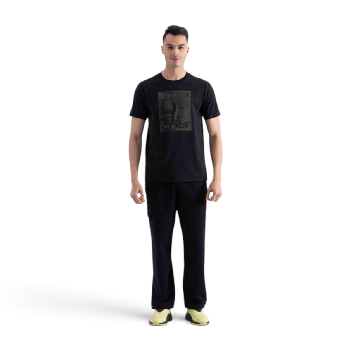 A man wearing a black FRG SS TEE with a graphic print on the chest, paired with black trousers and yellow sneakers.