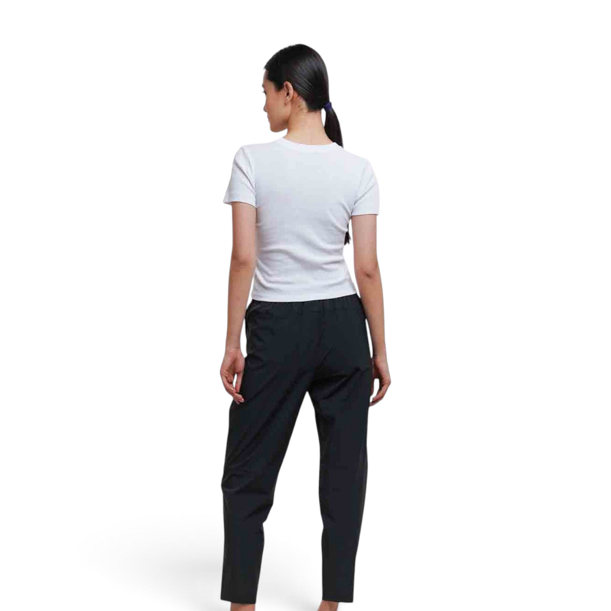 A woman wearing FRG black ankle pants and a white t-shirt, viewed from the back against a plain studio background.